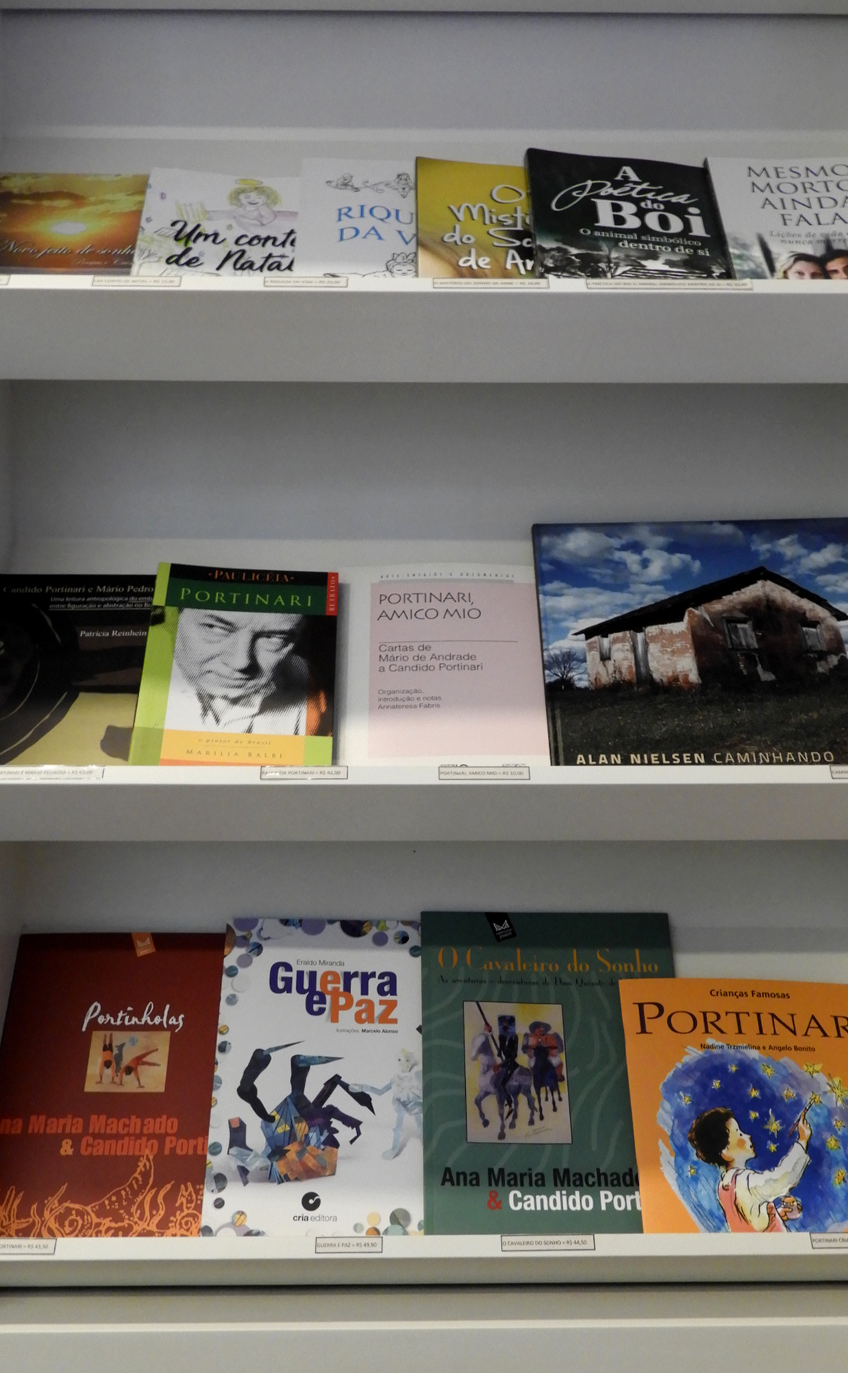 #PraCegoVer - Descrição da imagem para cegos: a primeira foto mostra em close alguns livros na prateleira da livraria. Em destaque obras sobre Portinari. A segunda imagem mostra uma estante branca com três nichos para livros e duas portas fechadas ao final do móvel. Dentro de cada nicho há vários livros à venda.