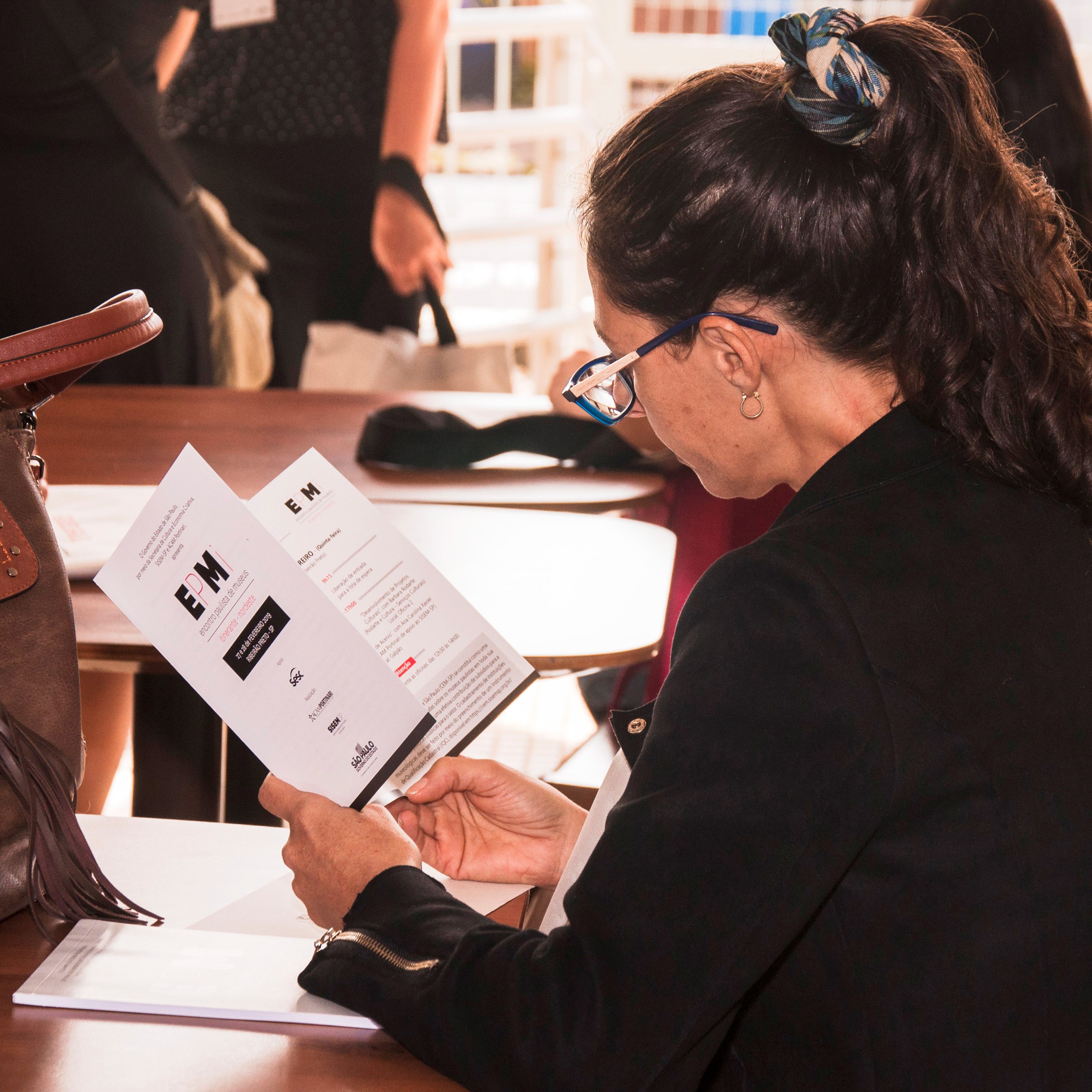 #PraCegoVer - Descrição da imagem para cegos: A foto mostra uma mulher de cabelos presos, usando óculos, lendo o um folder do EPMi.