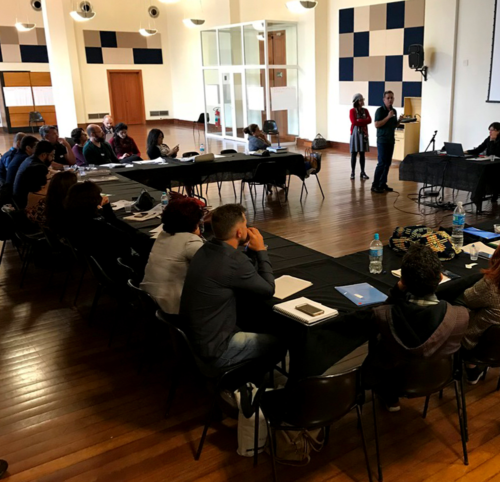 #PraCegoVer - Descrição da imagem para cegos: a foto mostra  um grupo de pessoas sentado numa mesa dentro de uma sala. Todos observam um telão à frente.