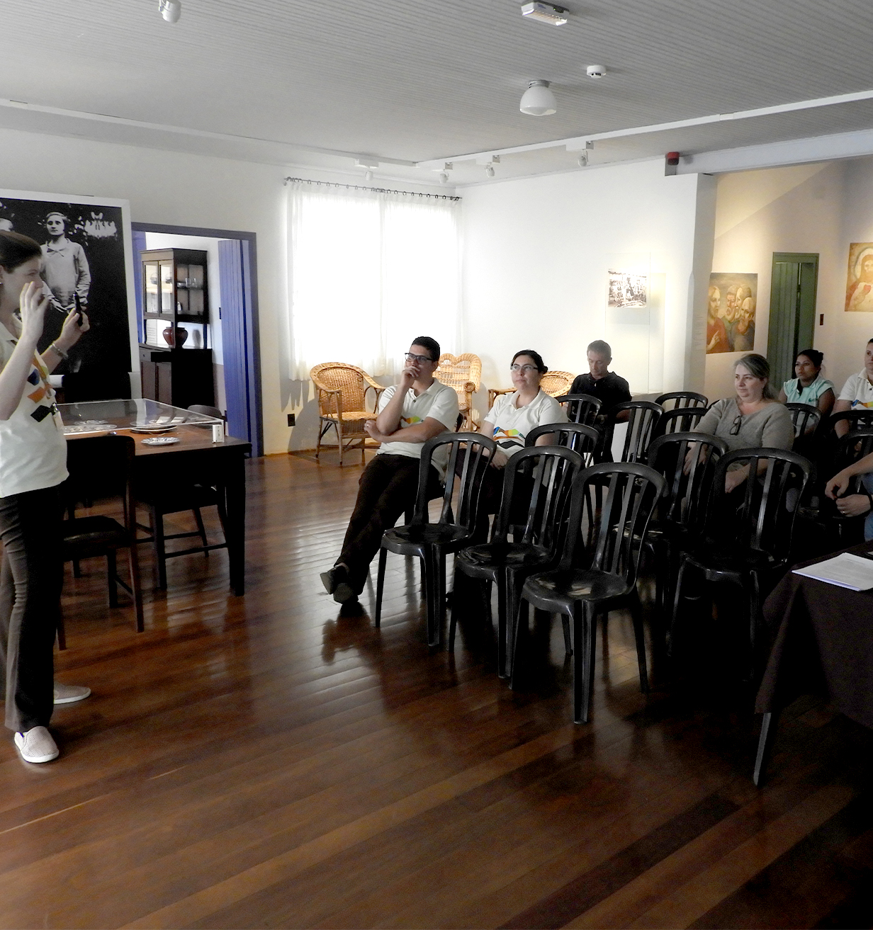 A foto mostra diversas pessoas sentadas na sala principal do Museu Casa de Portinari. Todos observam a palestrante que está à frente e em pé.