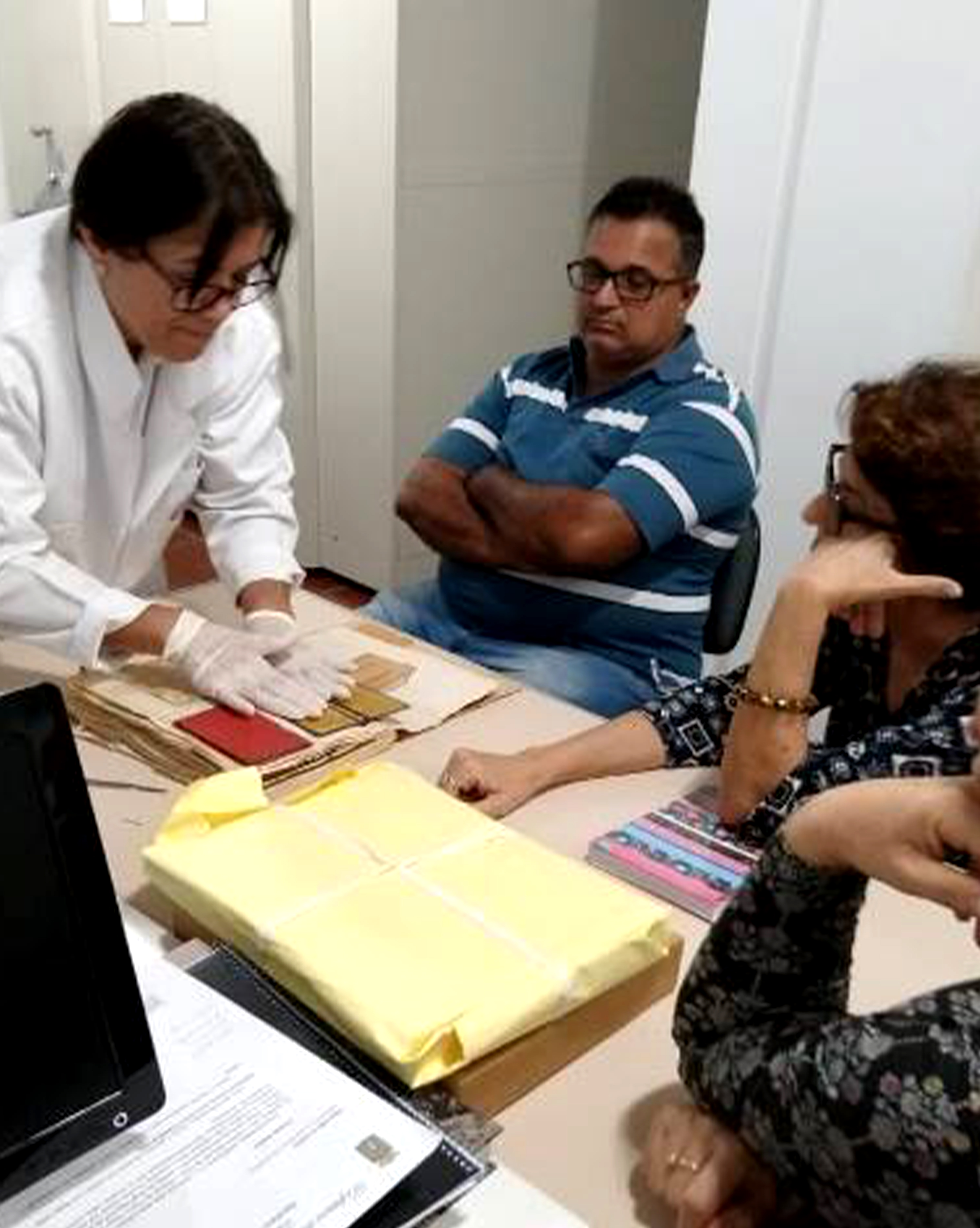 A imagem mostra três pessoas observando uma funcionária do museu manuseando papéis. A profissional está de jaleco branco e com luvas cirúrgicas. Todos estão ao redor de uma mesa, que tem diversos envelopes e um computador na superfície.