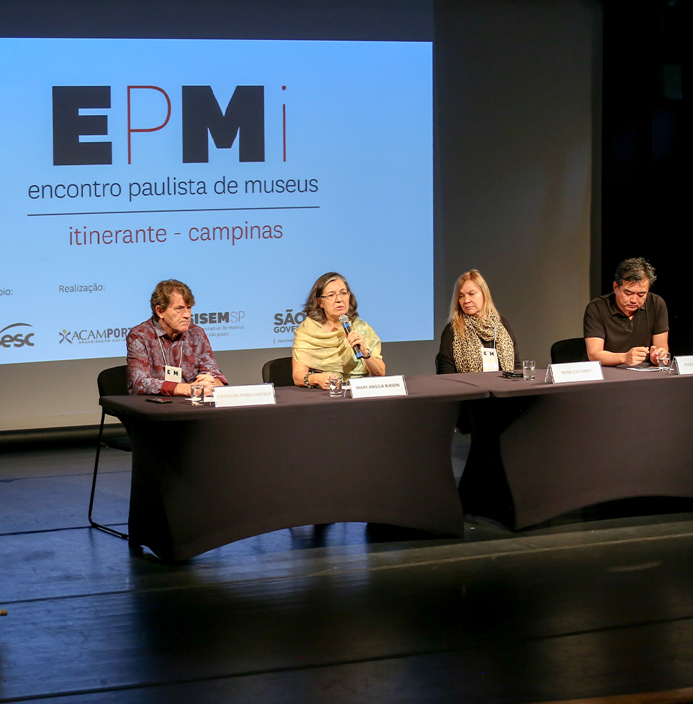 #PraCegoVer - Descrição da imagem para cegos: a foto mostra o palco do evento de
longe. No local aparecem um telão e um cartaz com os logos do EPMi. Ao centro está
a mesa com os participantes da mesa sentados.