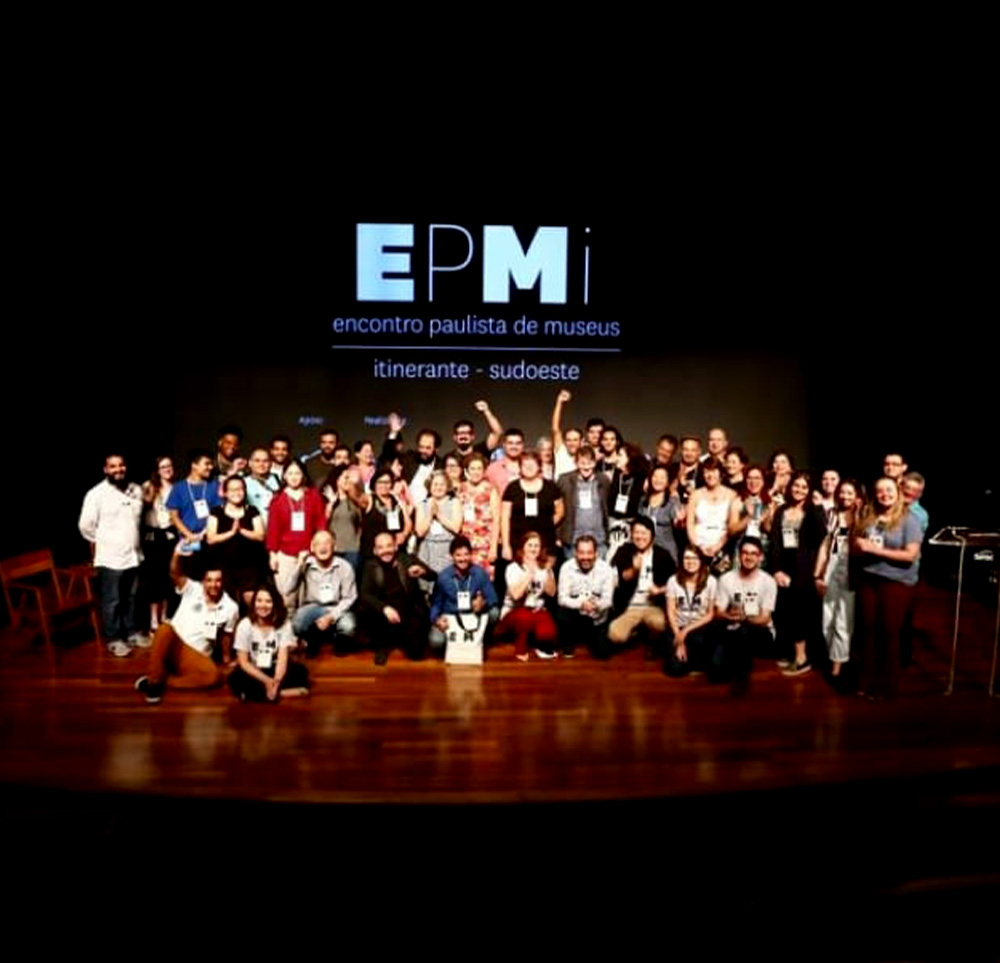 #PraCegoVer - Descrição da imagem para cegos: a imagem mostra um grupo de pessoas em cima de um palco posando para a foto, elas estão comemorando. Atrás delas aparece um telão com a marca “EPMi”.