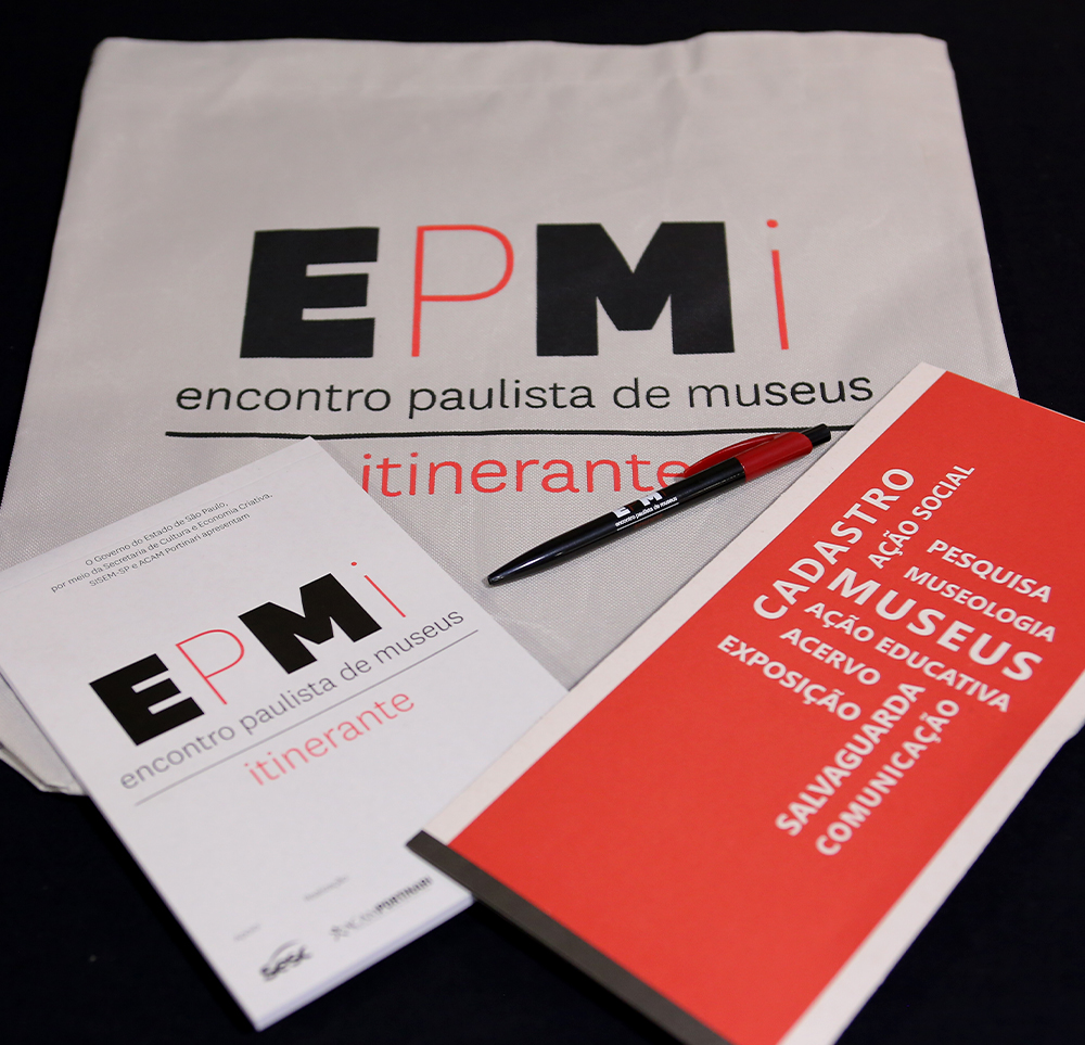 #PraCegoVer - Descrição da imagem para cegos: a imagem mostra um folder, um bloco de papel, uma caneta e uma sacola, todos com a logo do SISEM-SP, em cima de uma superfície preta.