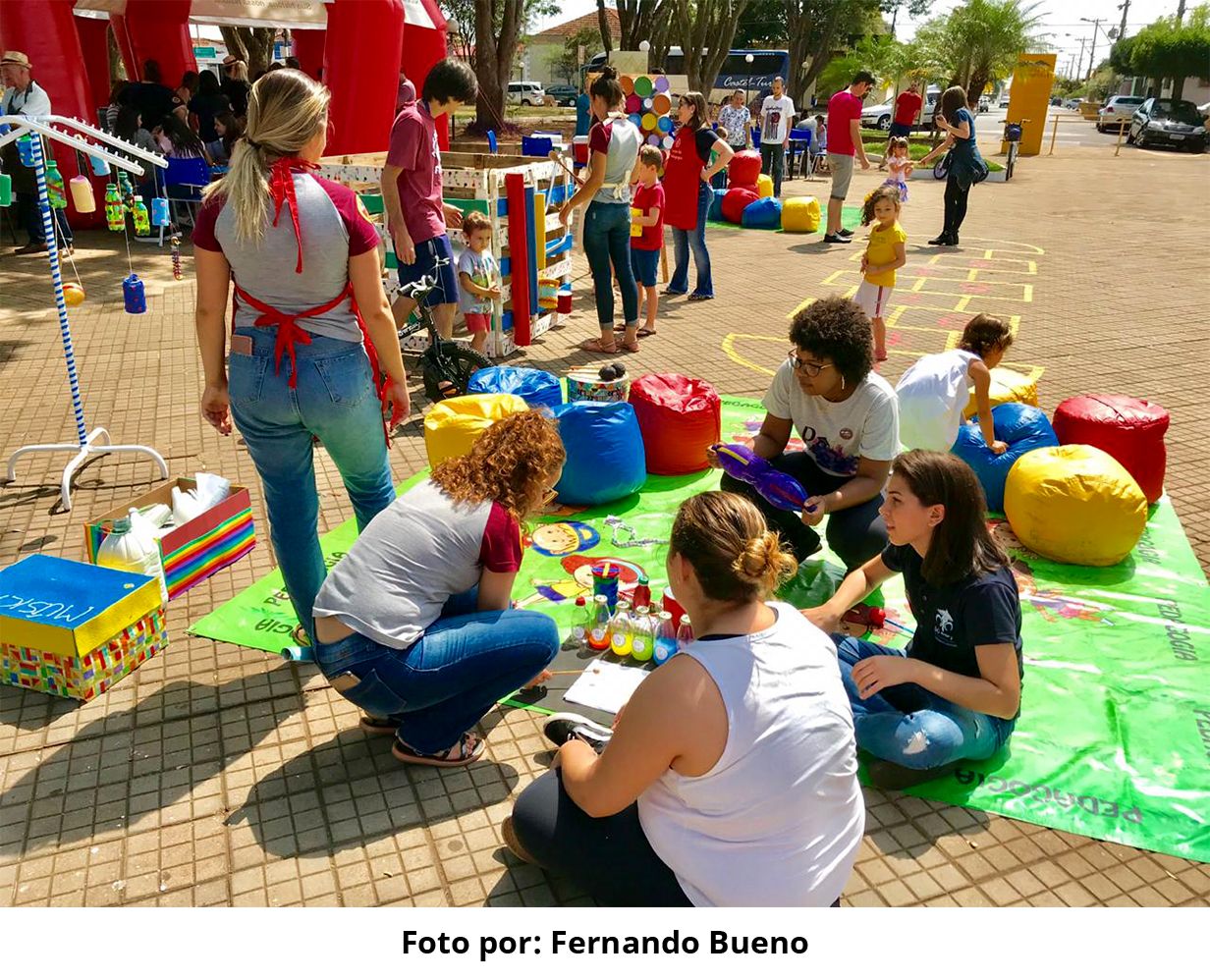 #PraCegoVer - Descrição da imagem para cegos: a foto mostra um grupo de pessoas, adultos e crianças, exercendo atividades na praça à frente ao Museu. Há vários brinquedos coloridos espalhados pelo local e um tapete verde, com pufes também coloridos no chão.