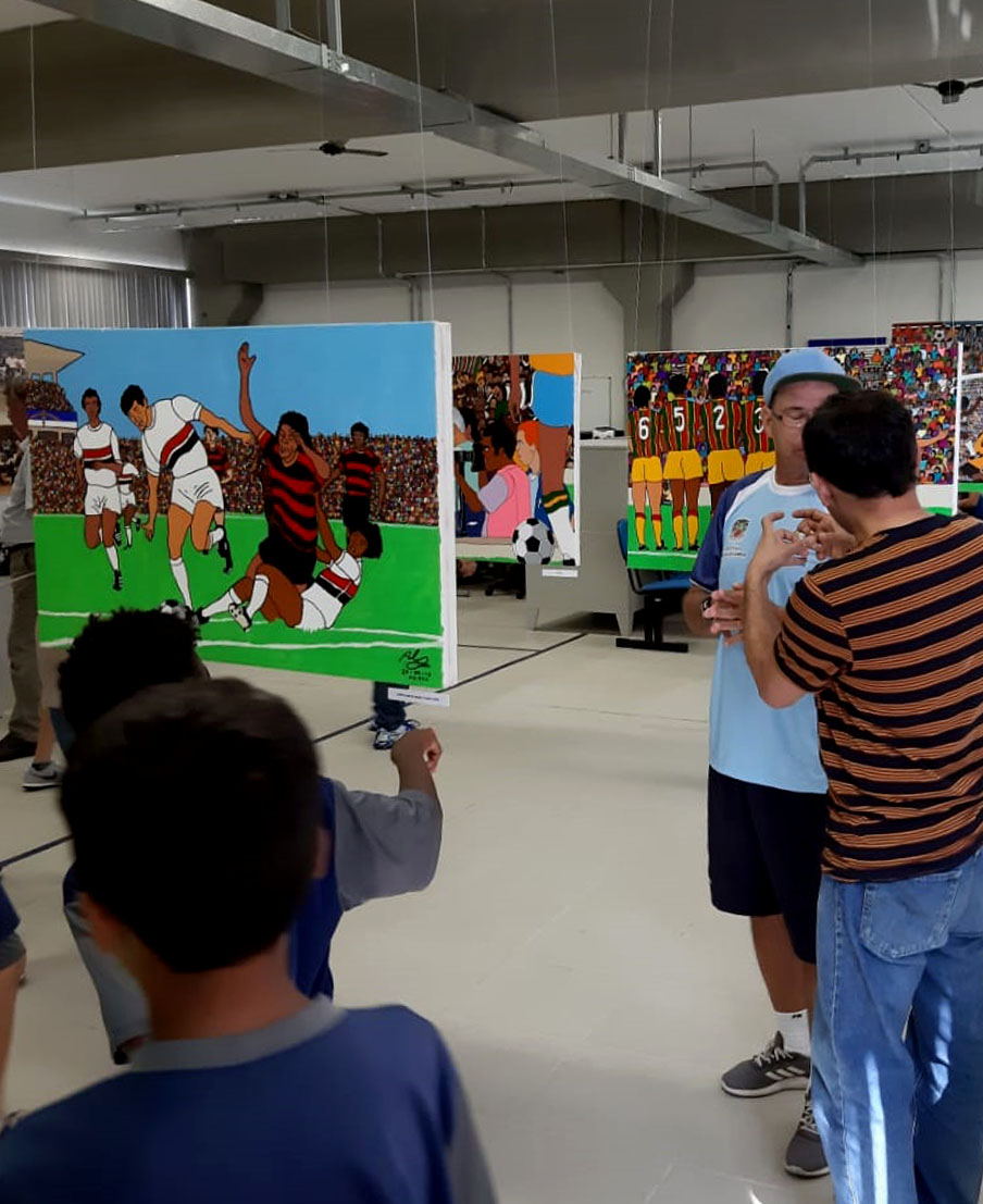 #PraCegoVer - Descrição da imagem para cegos: na imagem aparece um grupo de pessoas dentro de uma sala que contém diversas obras penduradas pelo teto. Nas peças aparecem ilustrações de jogadores de futebol.