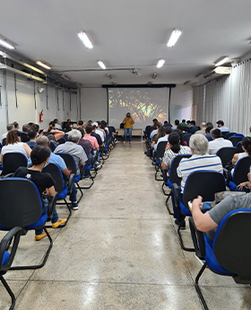 #PraTodosVerem – A imagem mostra a foto de uma sala com diversas pessoas sentadas assistindo a uma palestra. O fundo da fotografia exibe uma mulher em pé, uma mesa e um projetor com a foto de uma árvore na tela.