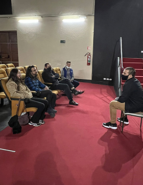 #PraTodosVerem – A imagem mostra um homem sentado conversando com quatro pessoas no Auditório Claudio Santoro. O funda mostra a lateral de uma escada e de uma porta.