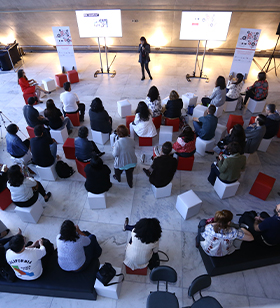 #PraTodosVerem – Foto de um grupo de pessoas em uma sala. Todos estão sentados assistindo a uma palestra ministrada por uma mulher.