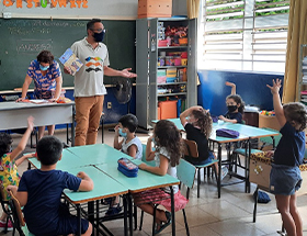 #PraTodosVerem – Imagem de uma sala de aula com crianças. Ao centro, existem dois grupos de mesas azuis com quatro alunos sentados em cada uma. Uma menina está em pé, com as mãos para cima, em frente ao educador do Museu Casa de Portinari. O fundo da sala mostra uma lousa verde, armário com livros e uma professora apoiando sobre uma mesa.