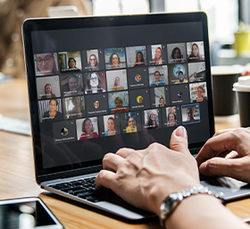 #PraTodosVerem – Foto de duas mãos mexendo em um notebook preto sobre uma mesa de madeira. A tela do aparelho exibe uma chamada de vídeo com diversas pessoas.