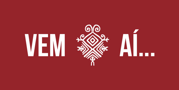 #PraCegoVer - Descrição da imagem para cegos: a imagem mostra, em um fundo vermelho, a frase: "Vem aí...". Entre a divisão das duas palavras, contém um símbolo indígena em branco que apresenta faixas diagonais, circulares e retas. Esse símbolo é o logo do novo Museu das Culturas Indígenas.