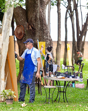 #PraCegoVer - Descrição da imagem para cegos: a imagem mostra, na lateral esquerda, uma área verde com três artistas pintando telas. Cada material está encostado em uma árvore. Ao centro da fotografia, cadeiras e uma mesa com objetos de pinturas compõem o fundo do local.