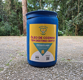 #PraCegoVer - Descrição da imagem para cegos: a imagem mostra, na lateral esquerda, um local arborizado. Ao centro da imagem, está um tambor azul, escrito: “Recicla Óleo. Óleo de cozinha tem destino certo. Deposite aqui”.
