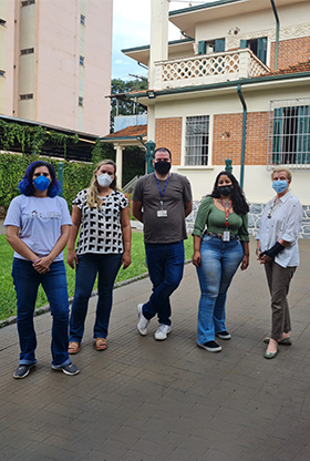#PraCegoVer - Descrição da imagem para cegos: a imagem mostra, na lateral esquerda, a fotografia de um grupo de cinco pessoas: um homem e quatro mulheres em pé, pousando para a foto. Eles estão no quintal de uma casa. A grama verde, janelas e uma sacada compõem o fundo da imagem.