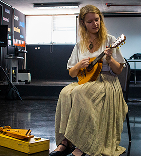 a imagem mostra, na lateral esquerda, a foto de uma mulher sentada e tocando um instrumento musical. Ela usa vestido longo e sapatilha preta. Ao lado dela, no chão, tem outros instrumentos musicais. Ao fundo, há uma parede com palavras escritas, uma mesa, um projetor e alguns equipamentos da sala do Museu Índia Vanuíre