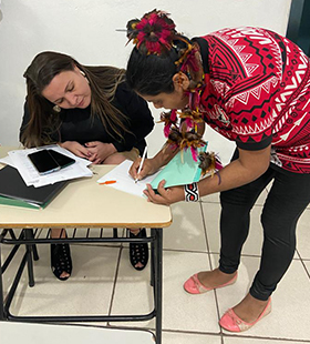 #PraTodosVerem – A imagem mostra, na lateral esquerda, a foto de uma indígena. Ela está abaixada sobre uma mesa escolar, assinando um papel, enquanto uma mulher, que está sentada na cadeira, observa a ação. Sobre a mesa, há alguns papéis e celulares. A indígena está usando um adorno de cabeça e vestindo camiseta vermelha com desenhos de traços indígenas.
