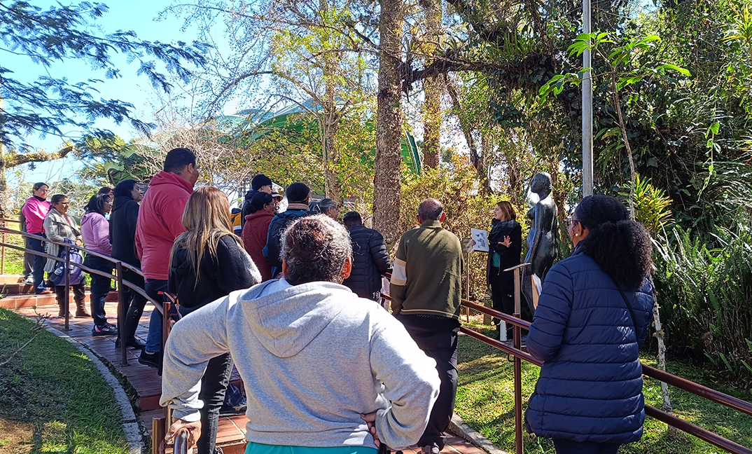 Grupo de visitantes de costas observando uma escultura de bronze em um jardim, durante visita guiada ao ar livre.