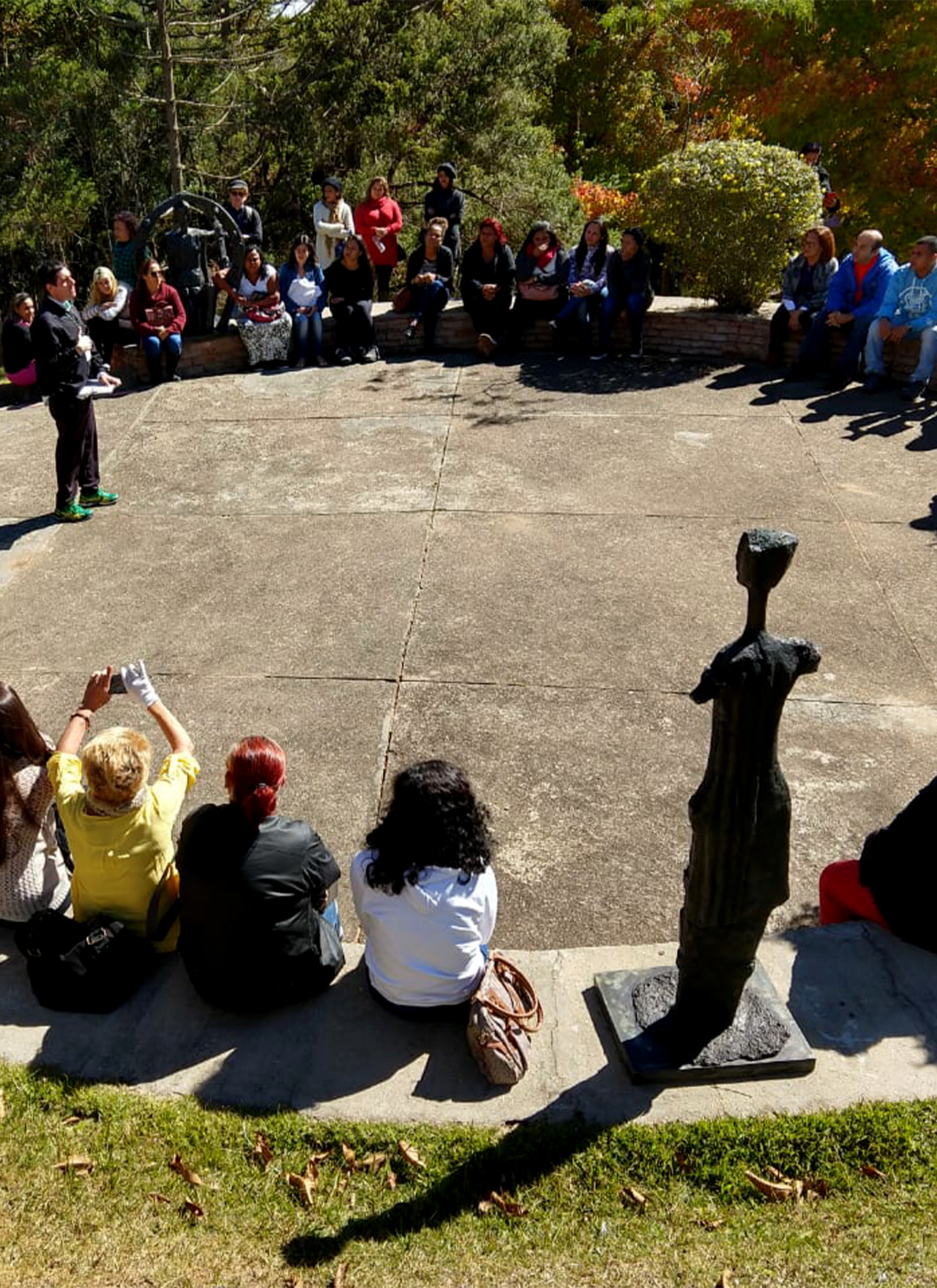 #PraCegoVer - Descrição da imagem para cegos: a foto mostra um grupo de pessoas sentado numa parte externa do Museu. São homens, mulheres, jovens e adultos. Eles estão em roda e todos observam o educador do equipamento, que está falando ao centro. O local é arborizado e algumas esculturas de Felícia Leirner aparecem no meio das pessoas.
