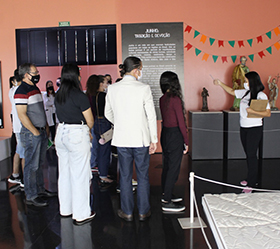 #PraTodosVerem –A imagem mostra um grupo de pessoas em uma sala expositiva do Museu Índia Vanuíre. Eles estão acompanhando a uma visita técnica realizada por uma educadora da instituição.