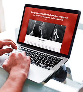 #PraCegoVer - Descrição da imagem para cegos: a imagem mostra, na lateral esquerda, a foto de duas mãos mexendo em um notebook que está aberto na exposição virtual do Museu Índia Vanuíre. A tela do aparelho mostra a fotografia de uma mulher indígena e um fundo vermelho.