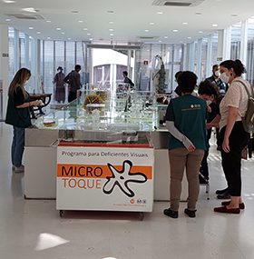 foto colorida mostra sala branca com paredes de vidro e, ao fundo, duas portas brancas. No centro da foto há um mostruário com artefatos de vidro e peças coloridas. Na frente dele, uma placa de fundo branco mostra o texto “Programa para deficientes visuais. Micro toque.” Ao redor do mostruário há nove pessoas, homens e mulheres, que caminham pela sala, fotografam a estrutura e conversam entre si. Todos estão com máscara cobrindo a boca e o nariz.