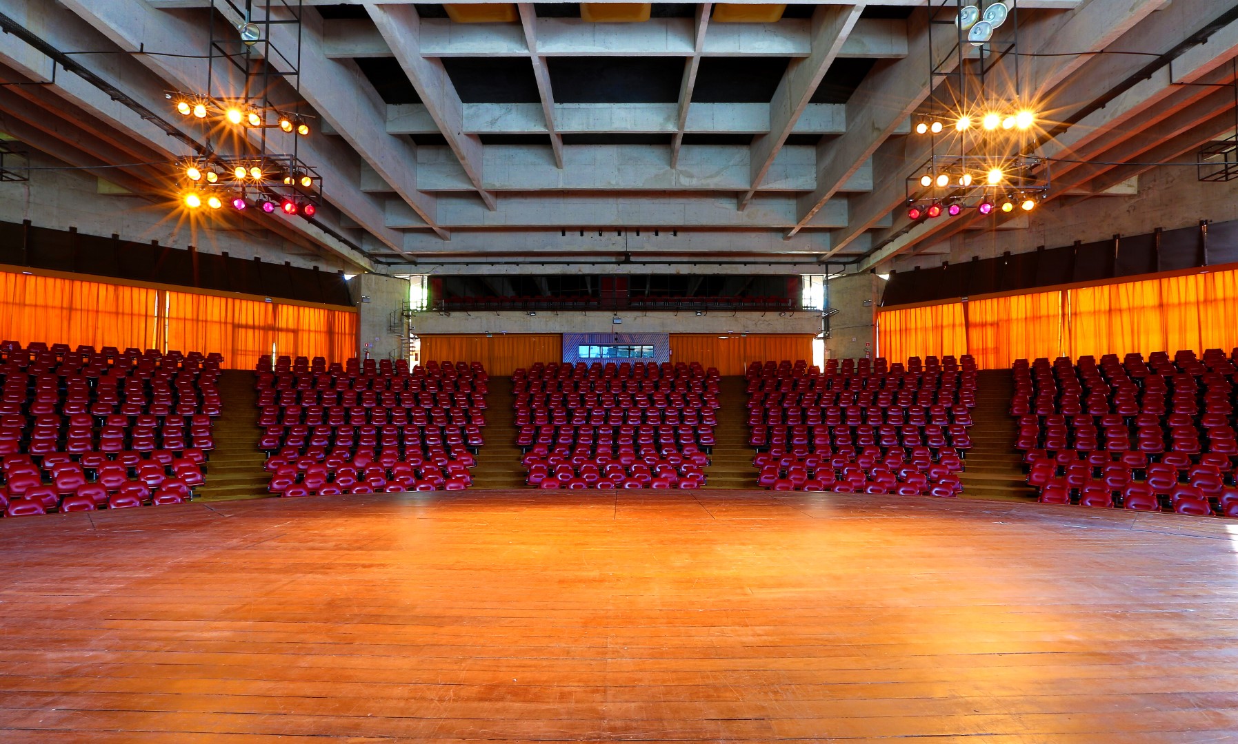 PraTodosVerem – A imagem mostra a visão do palco do Auditório Claudio Santoro vazio. O palco é de madeira e em frente exibe cinco conjuntos com fileiras de cadeiras vermelhas e acolchoadas. 