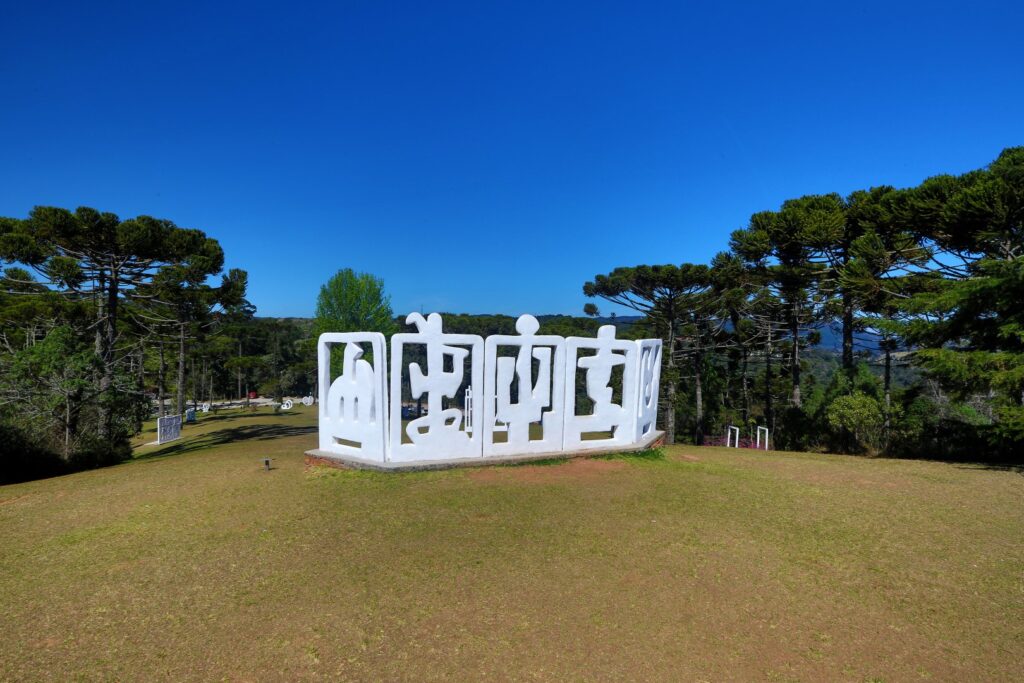 ParaTodosVerem: Gramado com escultura geométrica de concreto branca no centro. Ao redor, árvores verdes e céu azul. 