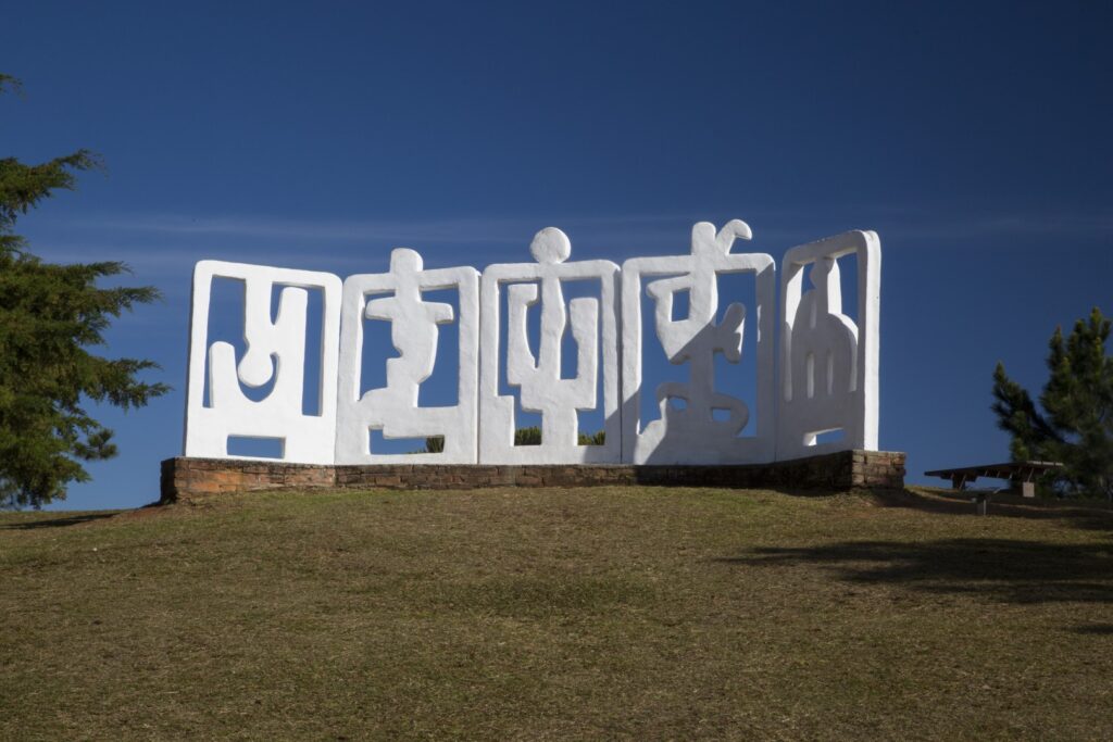 ParaTodosVerem: Gramado com escultura geométrica de concreto branca no centro. Ao redor, árvores verdes e céu azul. 