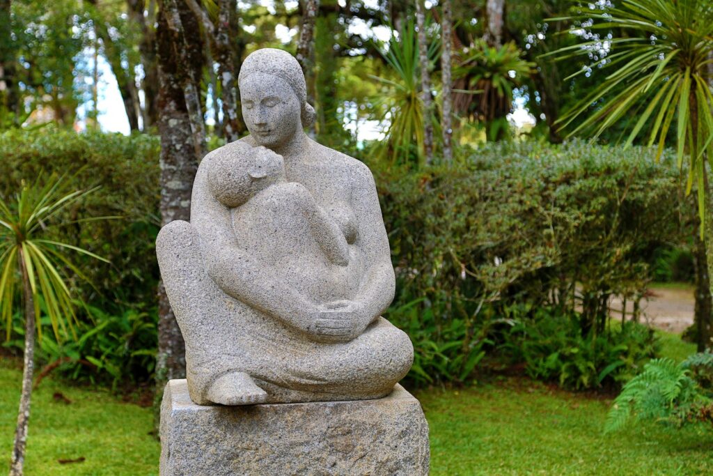 ParaTodosVerem: Escultura de concreto cinza de uma mulher sentada, abraçando uma criança. A escultura está apoiada em uma estrutura também de concreto. Ao fundo, arbustos e árvores verdes.