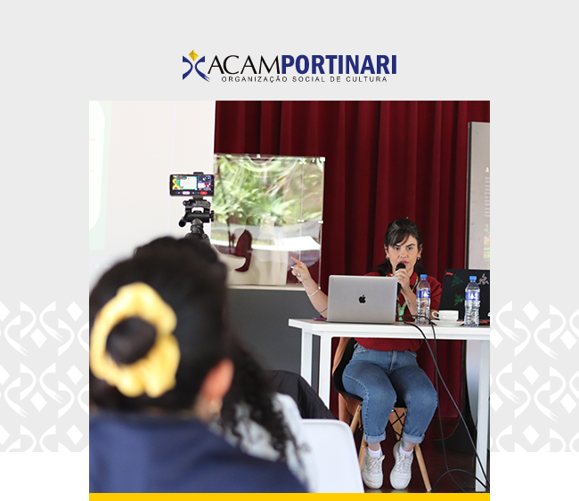 No alto da imagem, há um logotipo da ACAM Portinari nas cores azul e amarela, com o texto “ACAM Portinari” e a inscrição “Organização Social de Cultura”. Abaixo, uma mulher sentada atrás de uma mesa branca fala ao microfone enquanto gesticula com uma das mãos. Diante dela, pessoas assistem sentadas, de costas. Ao lado da mesa, uma câmera sobre tripé grava a cena. O ambiente é fechado, com cortinas vermelhas ao fundo e iluminação suave.
