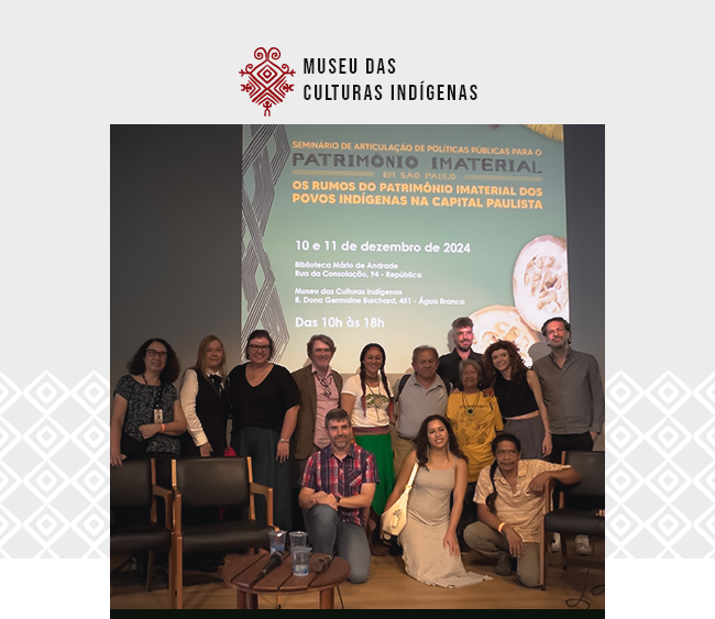na parte superior da imagem, está o logotipo do Museu das Culturas Indígenas. A cena mostra um grupo diverso de pessoas posando para uma foto em frente a um painel de divulgação de um seminário intitulado "Os Rumos do Patrimônio Imaterial dos Povos Indígenas na Capital Paulista", com datas e locais indicados para o evento. O painel possui tons de verde e amarelo, e contém ilustrações de artefatos indígenas. O grupo é composto por pessoas de diferentes idades, com expressões alegres e descontraídas. Algumas pessoas estão vestindo roupas tradicionais indígenas, enquanto outras vestem roupas casuais ou formais. Na frente, há duas cadeiras e uma pequena mesa com garrafas de água e papéis. Uma mulher agachada usa um vestido amarelo e sorri para a câmera. Ao lado dela, um homem de camisa xadrez vermelha está sentado no chão.