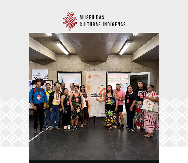Na parte superior da imagem, sobre fundo claro, aparece o logotipo do Museu das Culturas Indígenas. Ao centro, uma fotografia mostra um grupo de pessoas reunidas em um espaço interno, posando para a câmera. Os participantes vestem roupas variadas, algumas com elementos tradicionais e acessórios culturais, e utilizam crachás de identificação. Ao fundo, há portas abertas e um banner com a inscrição “Povos Indígenas” posicionado ao centro. O ambiente possui iluminação artificial, com luminárias no teto e paredes de concreto aparente. Nas laterais da composição, aparecem padrões gráficos discretos que dialogam com a identidade visual da instituição.