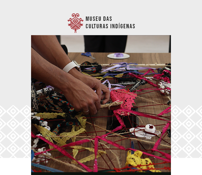 Imagem com fundo claro e padrões gráficos discretos nas laterais. Na parte superior, centralizado, está o logotipo do Museu das Culturas Indígenas em vermelho. Abaixo, uma fotografia mostra mãos trabalhando sobre uma mesa repleta de tiras de tecido coloridas, fitas e rendas em tons vibrantes de rosa, amarelo, preto e outros. As fitas se entrelaçam formando um emaranhado, sugerindo uma atividade coletiva de arte ou artesanato. Ao fundo, desfocados, aparecem outros materiais e ferramentas, como tesouras e rolos de fita, reforçando o ambiente criativo e colaborativo.