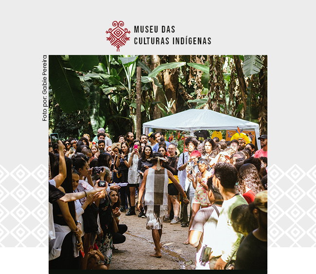 Na parte superior da imagem, sobre fundo claro, aparece o logotipo do Museu das Culturas Indígenas. Ao centro da composição, uma fotografia mostra um desfile realizado em área externa cercada por vegetação tropical, com bananeiras e outras plantas ao fundo. Uma pessoa indígena caminha pelo espaço central, usando vestimenta tradicional composta por tecido em tons naturais e adorno na cabeça. Nas laterais, um grande público forma um corredor humano, observando, fotografando e registrando o momento com celulares. Ao fundo, é possível ver uma tenda branca e outras pessoas acompanhando a atividade. A cena ocorre em ambiente iluminado por luz natural, sugerindo um evento cultural ao ar livre. Na lateral esquerda da imagem aparece a inscrição vertical Foto por: Gabie Pereira. Nas laterais da composição também aparecem padrões gráficos discretos que dialogam com a identidade visual da instituição.