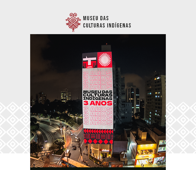 A imagem mostra um prédio alto em São Paulo iluminado à noite com uma grande projeção comemorativa na sua fachada. No centro da projeção está escrito em letras grandes: “Museu das Culturas Indígenas – 3 ANOS”. O número “3 ANOS” se repete várias vezes em vermelho e branco ao redor do texto principal. Acima da frase, aparecem símbolos gráficos indígenas em vermelho e branco, incluindo formas geométricas e um círculo estilizado que remete ao sol. Na parte inferior da projeção, há mais grafismos indígenas. A cena noturna mostra o entorno urbano iluminado por postes e faróis de carros em movimento. Acima da imagem, aparece o logotipo do Museu das Culturas Indígenas, com um símbolo gráfico vermelho e o nome da instituição escrito em letras pretas. A imagem celebra os três anos de funcionamento do museu dedicado às culturas indígenas, localizado na capital paulista.