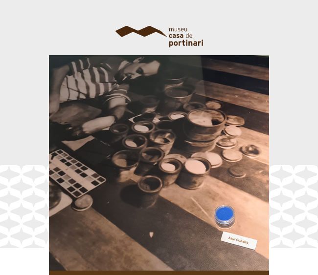 na parte superior da imagem, está o logotipo do Museu Casa de Portinari. Em destaque, uma fotografia em preto e branco retrata um homem, o pintor Candido Portinari, sentado no chão com uma expressão focada. Ele veste uma camisa listrada e calça clara, e está cercado por diversos potes de tinta, pincéis e uma paleta de cores. A fotografia mostra uma cena de trabalho artístico, com os potes organizados em uma superfície de madeira marcada por listras. Na parte inferior direita da imagem, sobre a fotografia, há um pequeno recipiente contendo pigmento azul intenso identificado como “Azul Cobalto”. Uma etiqueta branca com o mesmo nome está posicionada logo abaixo. O fundo da imagem é cinza claro com um padrão decorativo sutil nas laterais.