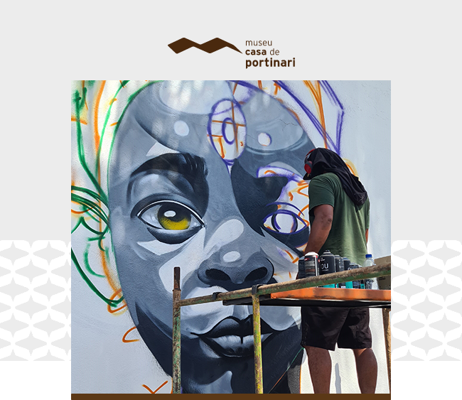 Imagem com o logotipo do Museu Casa de Portinari centralizado na parte superior, sobre fundo claro. Abaixo, um artista de camiseta verde, bermuda preta e rosto parcialmente coberto pinta um grande mural que retrata o rosto de uma pessoa negra com olhar expressivo. A obra, ainda em processo, combina tons de cinza, preto, branco, amarelo e detalhes em laranja e roxo, com traços e esboços visíveis. O artista está sobre um andaime, com latas de tinta spray dispostas à sua frente. Elementos gráficos da identidade visual aparecem nas laterais e na base da imagem.