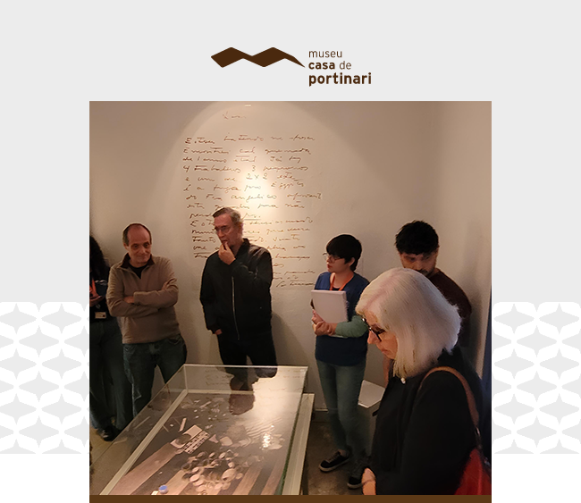 Imagem com fundo claro e detalhes gráficos discretos nas laterais. Na parte superior, centralizado, aparece o logotipo do Museu Casa de Portinari. Abaixo, uma fotografia mostra um grupo de seis pessoas reunido em uma sala expositiva do Museu. No centro da sala, há uma vitrine de vidro com fotografias e objetos expostos. Ao fundo, sobre a parede branca, vê-se um trecho de texto manuscrito em letras cursivas. As pessoas observam atentamente a vitrine e interagem entre si, transmitindo um momento de apreciação e reflexão sobre o acervo.