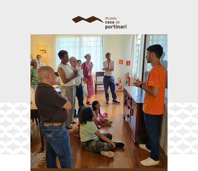 Em um ambiente interno do Museu Casa de Portinari, um grupo de pessoas participa de uma atividade mediada. À direita, um educador, de pé e vestindo camiseta laranja, fala e gesticula enquanto se dirige ao grupo. À frente dele, algumas pessoas estão sentadas no chão, incluindo uma criança, enquanto outras permanecem em pé, formando um semicírculo e observando atentamente. O espaço tem piso de madeira e iluminação natural vinda de janelas ao fundo, com cortinas claras. Ao centro, há um móvel antigo de madeira, que parece ser o foco da explicação. O ambiente é acolhedor e sugere uma visita guiada ou atividade educativa dentro do museu.