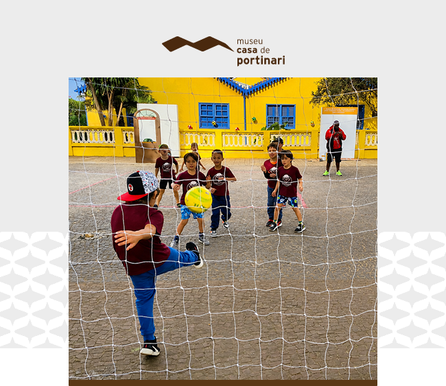 Na parte superior da imagem, sobre fundo cinza claro, aparece o logotipo do Museu Casa de Portinari. Ao centro, há uma fotografia em formato retangular. A imagem mostra um grupo de crianças em uma atividade de futebol realizada em área externa. Em primeiro plano, visto de costas, um menino veste camiseta vinho, calça azul e boné preto com detalhes vermelhos. Ele chuta uma bola amarela em direção ao gol. A rede ocupa grande parte da cena e aparece entre a câmera e os participantes. Do outro lado, várias crianças observam ou participam da brincadeira, distribuídas pela quadra. Ao fundo, destaca-se a fachada amarela da Casa de Portinari, com portas e janelas azuis, além de árvores e elementos do espaço externo. À direita da fotografia, uma pessoa adulta acompanha a atividade. Nas laterais da composição, sobre o fundo cinza, há padrões gráficos geométricos em tom branco. Na parte inferior da fotografia, uma faixa cinza acompanha a base da imagem.