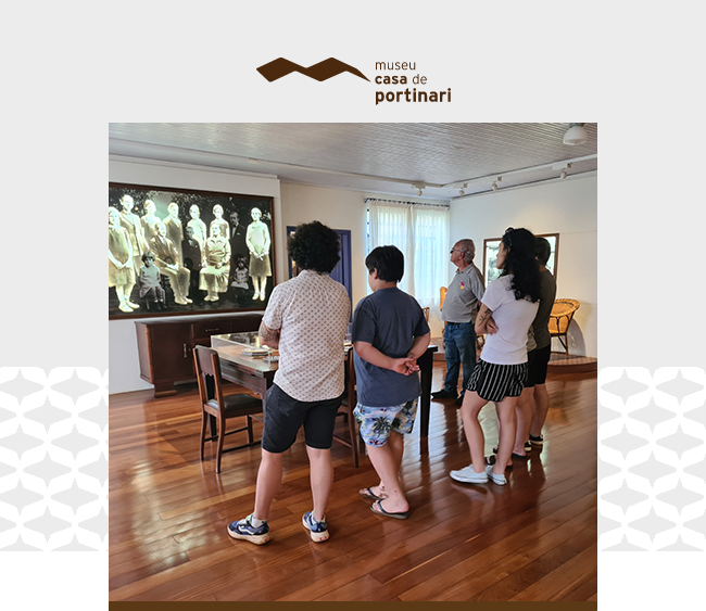 Na parte superior da imagem, o logotipo do Museu Casa de Portinari aparece centralizado sobre um fundo cinza claro, com elementos gráficos brancos nas laterais. Abaixo, a fotografia mostra um grupo de visitantes em um dos ambientes expositivos do museu. De costas, cinco pessoas observam atentamente uma fotografia em preto e branco de grandes dimensões que retrata a família de Portinari. O espaço tem piso de madeira, paredes claras e janelas que permitem a entrada de luz natural. À esquerda, uma mesa com cadeiras integra o ambiente de características domésticas.