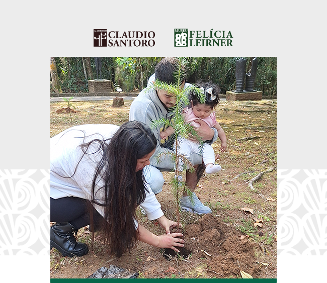 Na imagem, vemos uma família em um ambiente ao ar livre, em um jardim arborizado. Há três pessoas principais: uma mulher jovem que está ajoelhada no chão, plantando uma pequena muda de árvore; um homem jovem que segura uma criança no colo, sentado ao fundo, observando a cena. A criança, um bebê, também olha para a muda. O homem parece estar apoiando a mulher no processo de plantio, enquanto a criança está envolvida na atividade. O solo ao redor é marrom e úmido. Acima da foto, estão os logotipos do Museu Felícia Leirner e Auditório Claudio Santoro. A cena transmite uma mensagem de cuidado com o meio ambiente e conexão familiar.