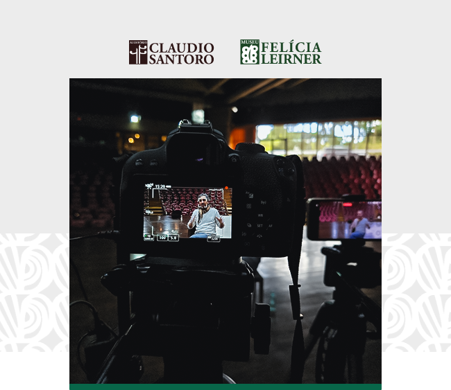 na parte superior da imagem, aparecem os logotipos do Auditório Claudio Santoro e do Museu Felícia Leirner. No centro, uma câmera profissional montada em um tripé está posicionada para gravar um homem sentado em uma cadeira no palco do auditório. O visor da câmera mostra o homem vestindo camisa branca e calça escura, gesticulando e falando diretamente para a câmera com uma expressão confiante. Ao fundo, fileiras de poltronas vermelhas estão dispostas no auditório, que está parcialmente iluminado por luzes suaves. À direita, um celular em um tripé grava a cena sob outro ângulo. O fundo da imagem é cinza claro com detalhes decorativos nas bordas inferiores.