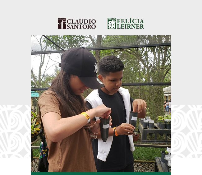 Na parte superior da imagem, sobre fundo claro, aparecem os logotipos do Auditório Claudio Santoro e do Museu Felícia Leirner. Ao centro, uma fotografia mostra duas crianças em um ambiente externo, cercado por vegetação. À esquerda, uma menina usa boné preto e camiseta marrom; ao lado, um menino veste camiseta preta com um casaco branco. Ambos seguram pequenos recipientes com terra, concentrados na atividade. Ao redor, há bancadas com mudas de plantas organizadas em vasos e bandejas. Ao fundo, árvores e estruturas do espaço expositivo ao ar livre compõem o cenário. Nas laterais da composição, aparecem padrões gráficos discretos que dialogam com a identidade visual da instituição.
