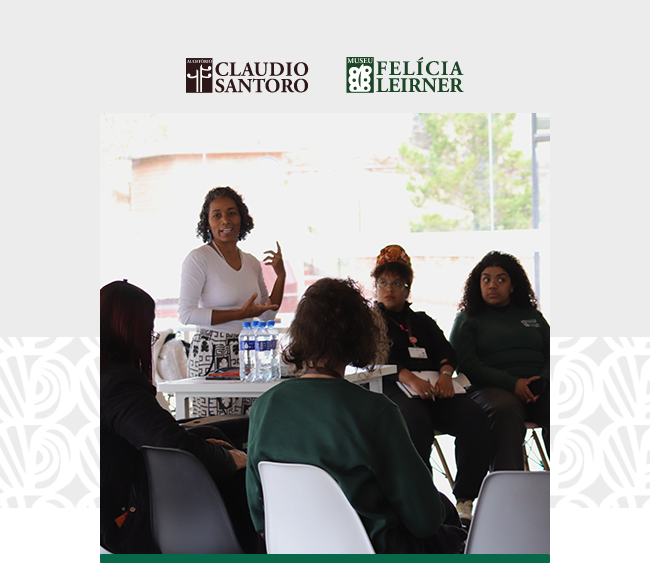 Imagem com os logotipos do Auditório Claudio Santoro e do Museu Felícia Leirner centralizados na parte superior, sobre fundo claro com grafismos discretos nas laterais. Abaixo, a fotografia principal mostra uma roda de conversa em ambiente iluminado por luz natural. Em pé, uma mulher fala ao grupo, gesticulando enquanto apresenta uma ideia. Sentadas ao redor da mesa, outras participantes acompanham atentamente; algumas vestem uniformes verdes. Sobre a mesa, garrafas de água e materiais de apoio indicam o caráter formativo do encontro. Ao fundo, grandes janelas revelam uma área externa arborizada.
