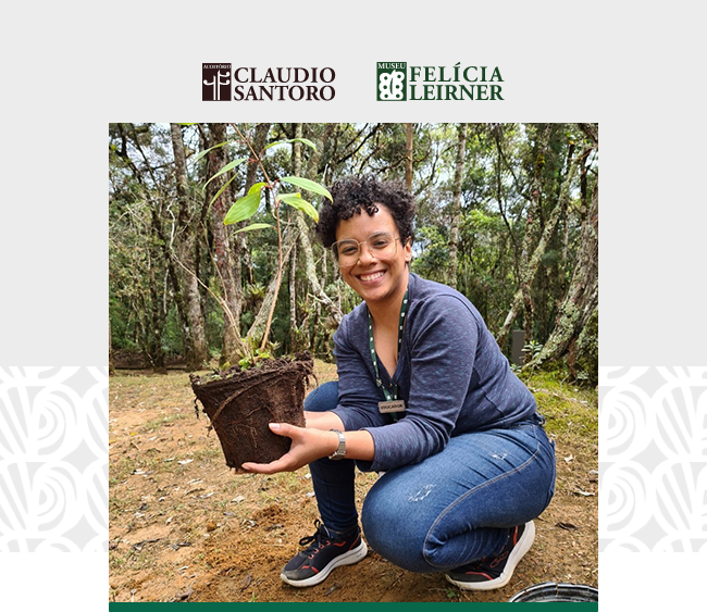 Na parte superior da imagem, sobre fundo claro, aparecem os logotipos do Auditório Claudio Santoro e do Museu Felícia Leirner. Ao centro da composição, uma fotografia mostra uma mulher agachada em área externa de mata, sorrindo para a câmera enquanto segura com as duas mãos uma muda de árvore em um pequeno recipiente com terra. Ela usa óculos de armação clara, blusa de manga longa em tom escuro, calça jeans e tênis. No peito, porta um crachá preso por cordão. Ao fundo, vê-se vegetação composta por árvores altas, folhas verdes e troncos cobertos por musgos, sugerindo ambiente natural de bosque ou parque. O solo é de terra batida, com pequenas pedras e folhas secas espalhadas. Nas laterais da composição aparecem padrões gráficos discretos que dialogam com a identidade visual das instituições.