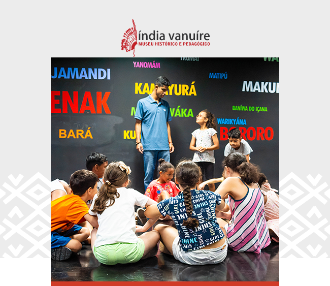 Imagem com o logotipo do Museu Índia Vanuíre centralizado na parte superior, sobre fundo claro. Abaixo, um grupo de crianças está reunido em roda no chão, em um espaço interno do museu. Ao fundo, um educador convidado conversa com duas crianças em pé. A parede atrás deles exibe nomes de povos indígenas escritos em letras grandes e coloridas, como "Yanomâmi", "Bororo", "Kamakã", "Makuráp", entre outros. Elementos gráficos com padrão indígena aparecem nas laterais da imagem.