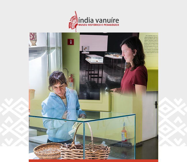Imagem com o logotipo do Museu Histórico e Pedagógico Índia Vanuíre centralizado na parte superior, sobre fundo claro com grafismos nas laterais. Abaixo, uma montagem de fotos de duas mulheres que observam vitrines de exposição no interior do museu. À esquerda, uma delas, vestindo jaqueta azul, observa atentamente um cesto de fibras naturais e objetos expostos. À direita, outra visitante, com blusa vermelha, analisa uma vitrine iluminada que contém peças e textos explicativos. O ambiente apresenta paredes em tons claros e iluminação direcionada às peças.