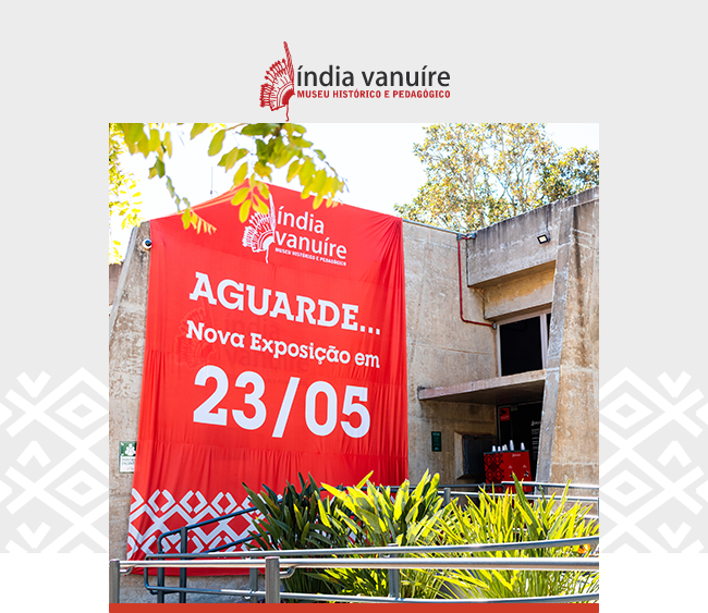 Na parte superior da imagem, sobre fundo cinza claro, aparece o logotipo do Museu Índia Vanuíre. Ao centro, uma fotografia mostra a área externa do museu em um dia ensolarado. Em destaque, uma grande faixa vermelha instalada na fachada traz a mensagem “AGUARDE... Nova Exposição em 23/05” em letras brancas. A estrutura do prédio aparece ao fundo, com paredes de concreto aparente e entrada parcialmente visível. Em primeiro plano, plantas verdes e corrimãos metálicos ocupam a parte inferior da imagem. Árvores com folhas iluminadas pela luz natural aparecem na parte superior esquerda. Nas laterais inferiores da composição, grafismos indígenas em branco aparecem sobre a faixa cinza de fundo.